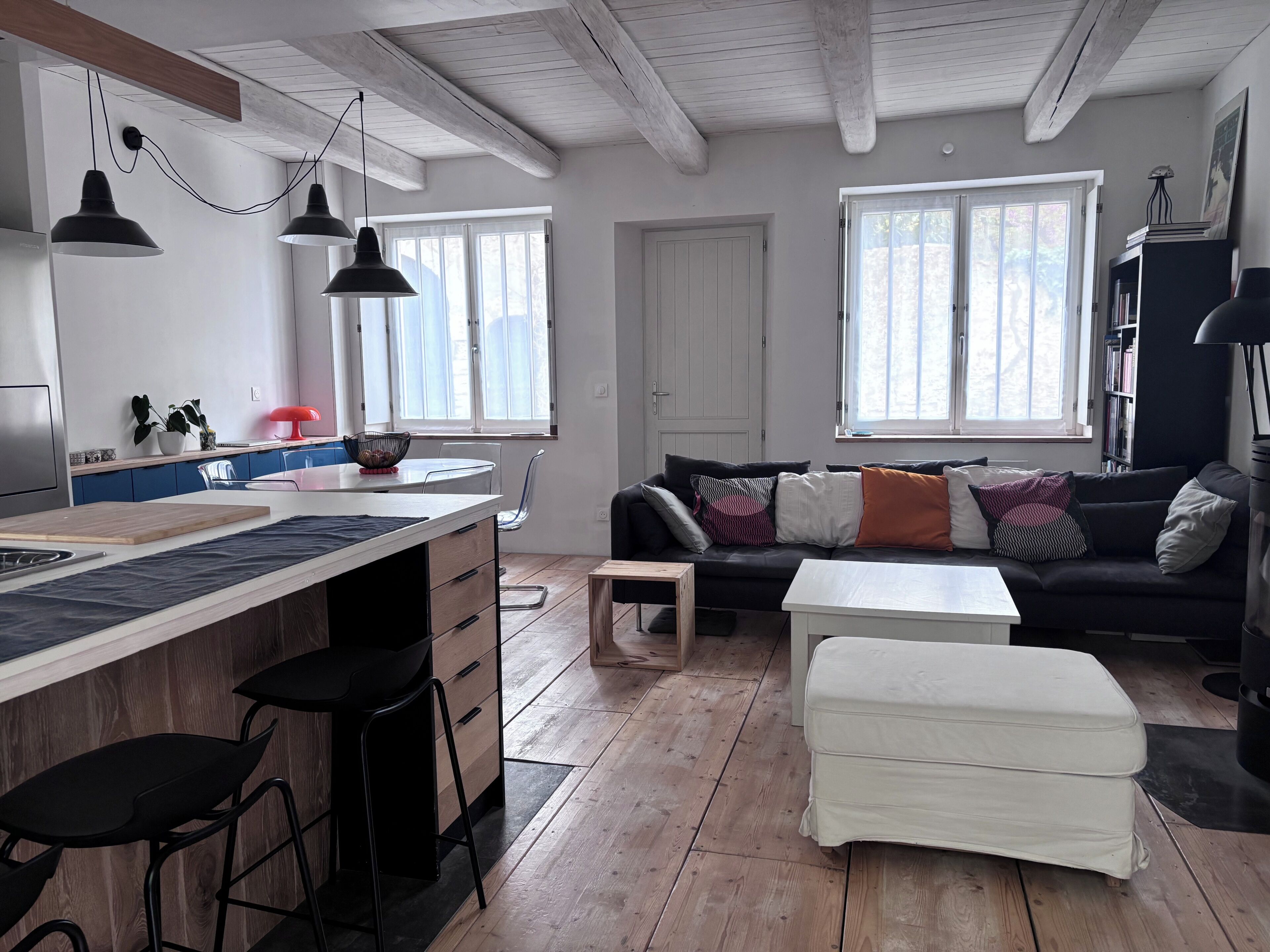 Et moderne køkken med en central ø, barstole, et spisebord og en sofa i en stue med store vinduer.