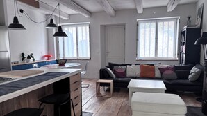Ett modernt kök med en central köksö, barstolar, ett matbord och en soffa i ett vardagsrum med stora fönster.