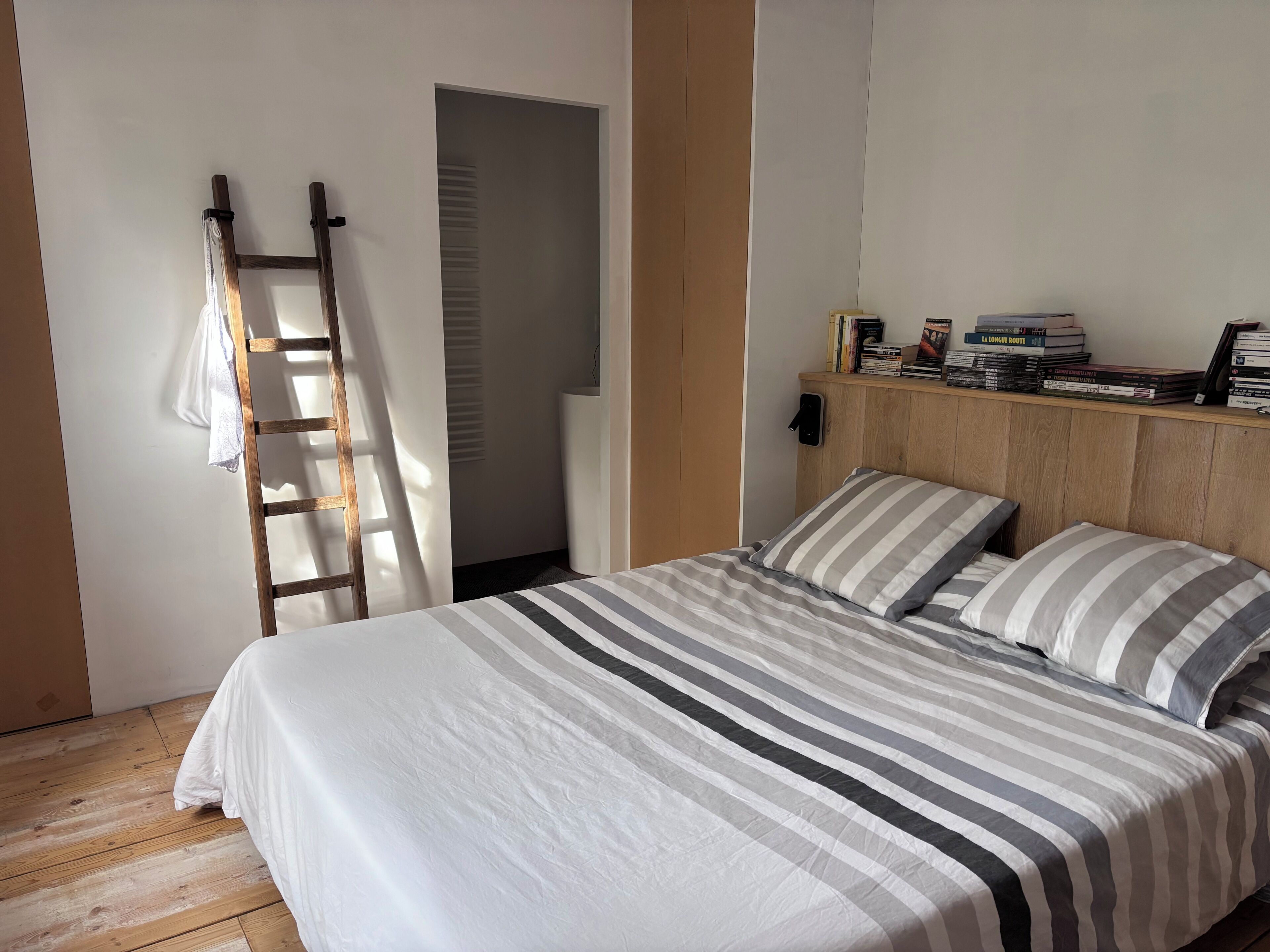 A bedroom with a bed, pillows, a wooden headboard, a bookshelf with books, a ladder, and a door leading to another room.