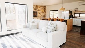 A modern living room with a white sofa, a stone accent wall, a dining area with wooden chairs, and a kitchen with white cabinets. - Sparrow House and Carolina Cottage- 2 lakeview homes, Sleeps 18 (Garfield)