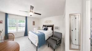 Une chambre d’hôtel avec un lit, une chaise, une table, un ventilateur de plafond et une fenêtre avec des rideaux.