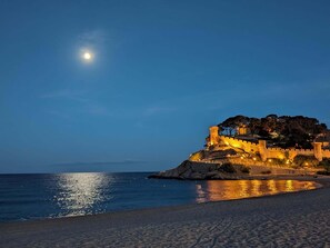 A coastal castle with a moat, a beach in the foreground, and a clear night sky with a visible moon. - EVH106 Apartment in the Heart of Tossa de Mar (Tossa de Mar)