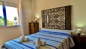 A hotel room with a bed, a window with curtains, a wooden dresser, a wall-mounted decorative panel, and a nightstand with a lamp. - EVH114 Terraced House with Communal Areas (Altafulla)