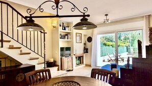 A dining area with a round table, chairs, a chandelier, a staircase, and a view through a glass door to an outdoor seating area. - EVH114 Terraced House with Communal Areas (Altafulla)