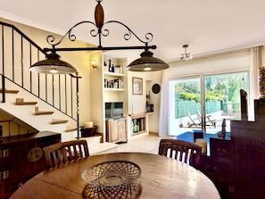 A dining area with a round table, chairs, a chandelier, a staircase, and a view through a glass door to an outdoor seating area.