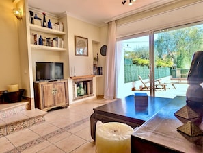 A living room with a TV, shelves with books and decorative items, a fireplace, and a view of the outdoors.