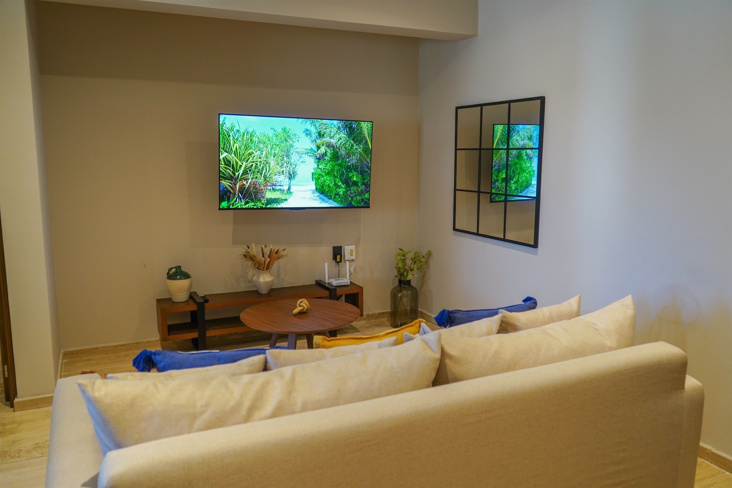 A living room with a flat-screen TV mounted on the wall, a round wooden coffee table, a beige sofa, and a small side table with decorative items.