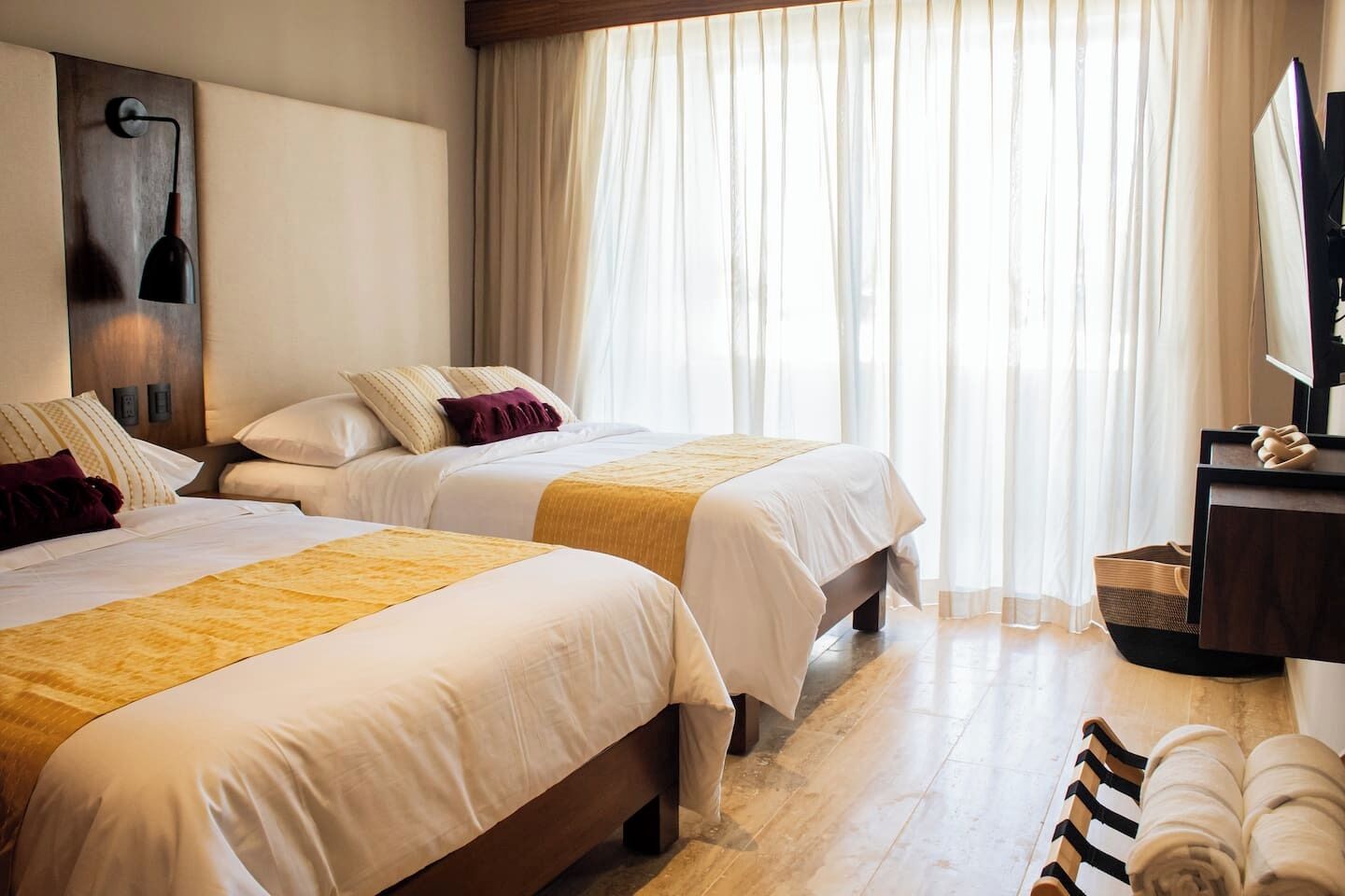 A hotel room with two beds, a large window with sheer curtains, a wall-mounted lamp, and a wooden floor.
