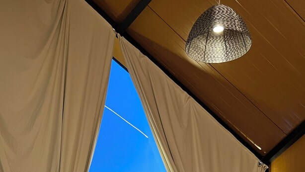 Een kamer met een groot raam, gordijnen, een hanglamp, een massagetafel en uitzicht op het omliggende landschap.