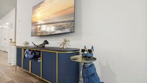 Ein modernes Interieur mit einem großen, an der Wand montierten Fernseher, der einen Sonnenuntergang über dem Meer zeigt, einem blau-goldenen Konsolentisch und einem kleinen runden Tisch mit einer blauen Tierfigur.
