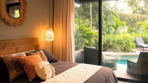 Ein Schlafzimmer mit einem großen Bett, einem runden Spiegel, einer Pendelleuchte und Blick auf einen Pool und Grünflächen.