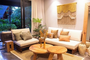 A living room with a sofa, armchair, coffee table, and a large window overlooking a garden. - SwimUp Retreat in Aldea Zama Pool at Your Door & Beach Club (Tulum)