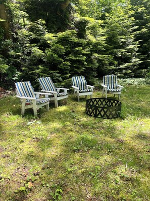 Drei Adirondack-Stühle mit gestreifter Polsterung in einer grünen Außenanlage, umgeben von dichten Bäumen.