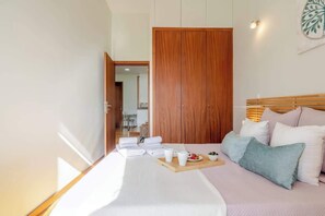 Ein Schlafzimmer mit einem Bett, Kissen, einem Nachttisch, einem Holzschrank und Blick in den angrenzenden Raum.