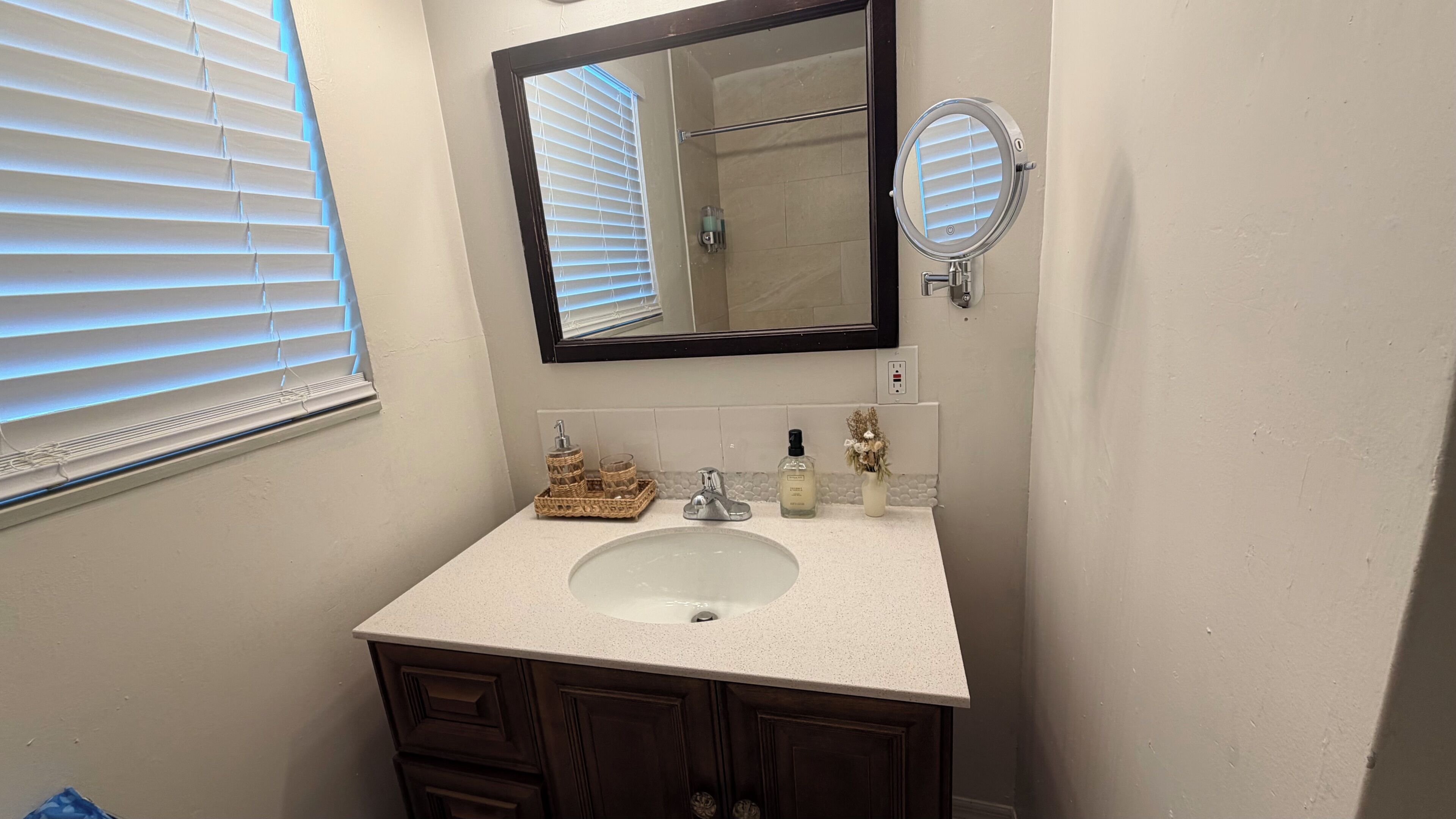 A bathroom with a white sink, dark wooden cabinets, a mirror, a small round magnifying mirror, and a window with blinds.