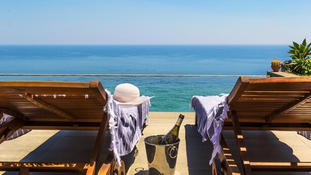 Un fauteuil de jardin en bois, avec une bouteille de champagne et un chapeau posés dessus, donnant sur une mer d’un bleu limpide.