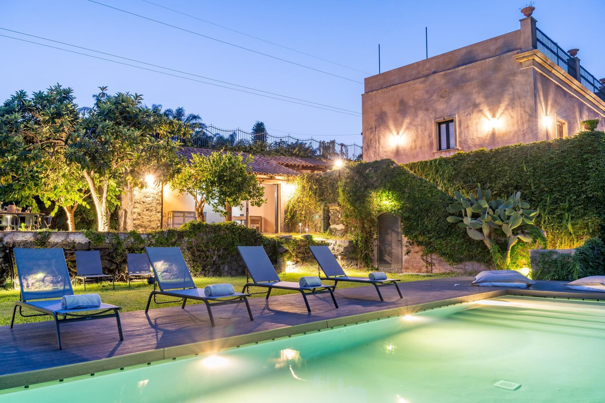 Un área junto a la piscina con sillones de descanso, piscina y un edificio con muros de piedra y techo de tejas.