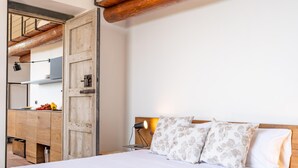 A bedroom with a wooden headboard, a bed with floral pillows, a nightstand with a lamp, and a view of a kitchen area. - Villa Tiaréa - Giarre (Giarre)