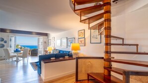 Una sala de estar moderna con una escalera de madera, un sofá, una zona de comedor y vista al mar.