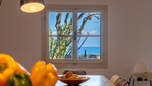 Uma sala de jantar com mesa, uma tigela de frutas, uma cadeirinha de bebê e uma janela com vista para o mar.