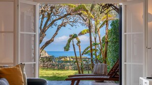 Quarto com uma grande janela, cama, um banco de madeira e vista para o oceano e vegetação.