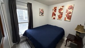 Ein Schlafzimmer mit Bett, einem Fenster mit Vorhängen, einem Nachttisch mit Lampe und floralen Bildern an der Wand.