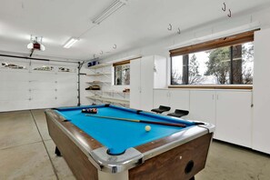 Ein Billardtisch mit blauer Spielfläche, eine weiße Kommode mit Regalen und ein Fenster mit Blick auf Bäume.