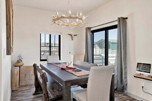 Uma sala de jantar com uma mesa de madeira, cadeiras brancas, um candelabro e uma janela ampla com vista.