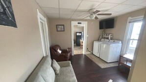 Ein Wohnzimmer mit einer Couch, einem braunen Ledersessel, einem Deckenventilator, einem Fernseher und einem Waschbereich mit einer Waschmaschine.