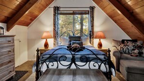 A bedroom with a sloped ceiling, a bed with a metal frame, two bedside lamps, a dresser, and a window with curtains. - Oakwood Cottage #2582 (Sugarloaf)