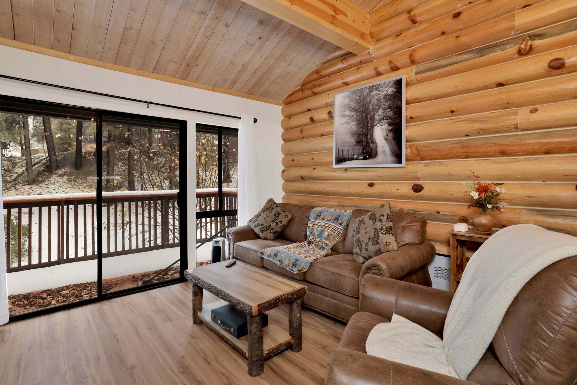 Interior de una cabaña de madera con sofá, mesa de centro y un ventanal con vistas a un bosque nevado.