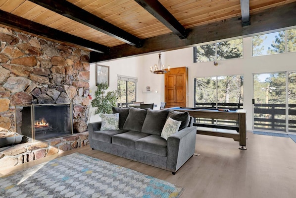 Ein gemütliches Wohnzimmer mit Kamin aus Stein, einem grauen Sofa, einem Billardtisch und Blick ins Freie.