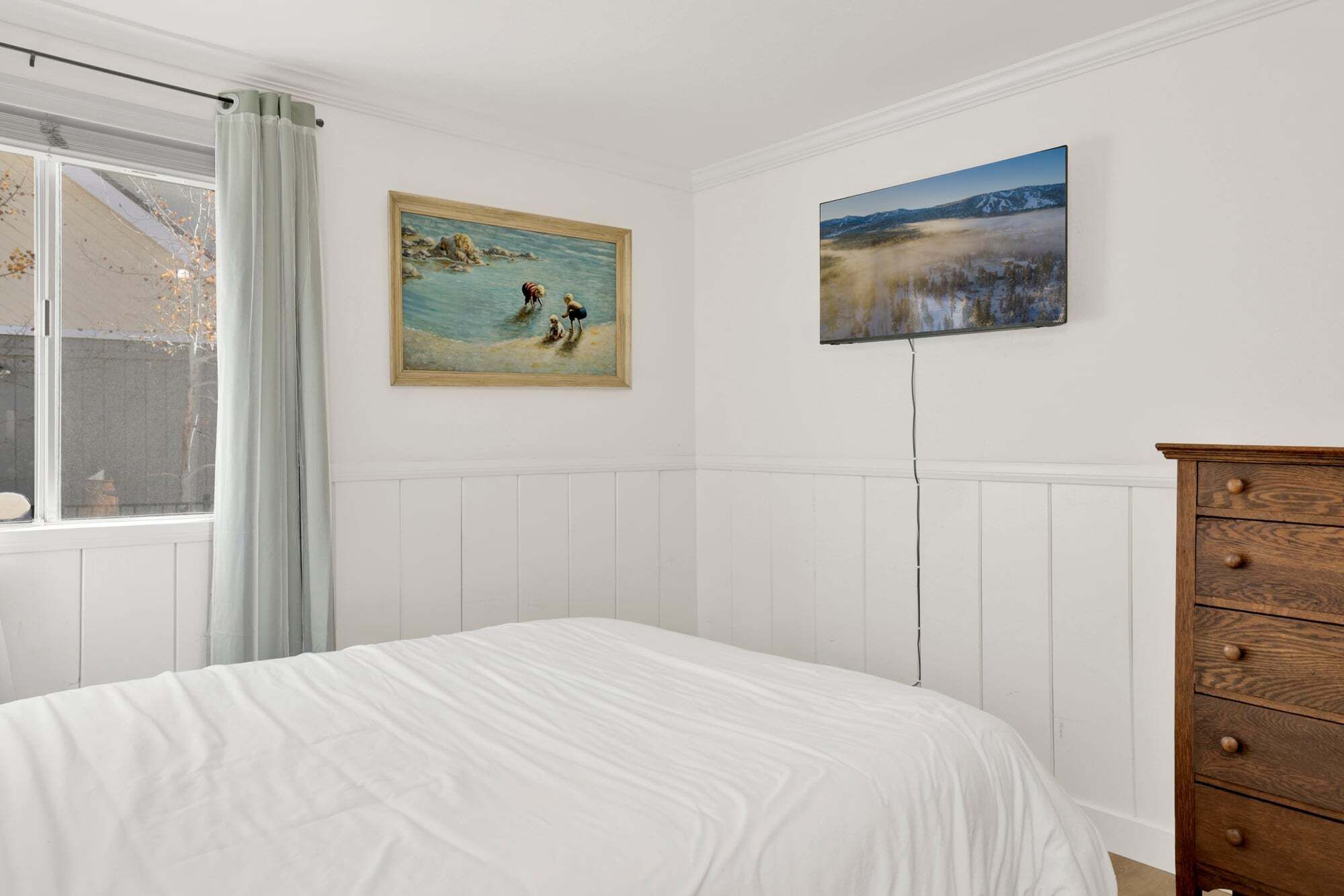 Un dormitorio con cama, cómoda de madera, ventana con cortinas y dos cuadros en la pared.