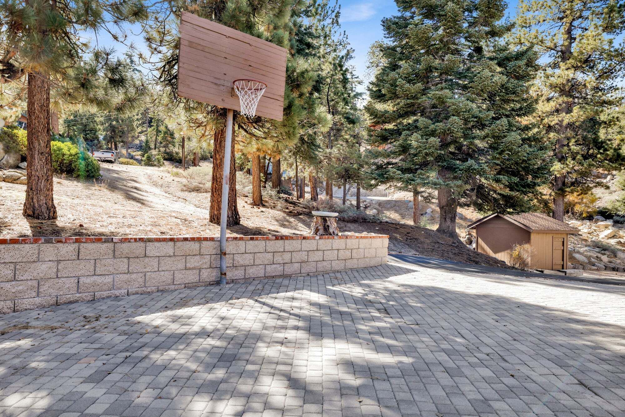A basketball court with a wooden backboard and hoop, surrounded by trees and a brick wall.