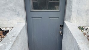 Una puerta gris con un pequeño ventanal arriba, un timbre metálico y una base de hormigón.