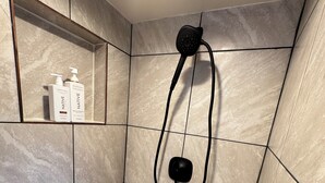 Zona de ducha con cabezal de ducha de mano, grifo empotrado en la pared y repisa con botellas de artículos de aseo.