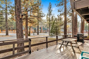 A wooden deck with a grill and picnic table, surrounded by tall pine trees. - 2594-willow Bear Lodge (Big Bear Lake)