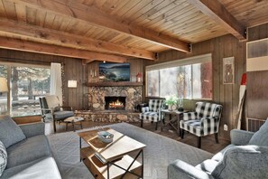 A cozy cabin interior with a stone fireplace, plaid chairs, and a large window overlooking a forest. - 2594-willow Bear Lodge (Big Bear Lake)