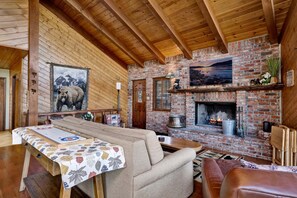A cozy room with a fireplace, wooden ceiling beams, a table with a leaf-patterned tablecloth, and a sofa. - 2600-the Tree Top Cabin (Big Bear Lake)