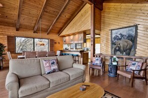 A cozy cabin interior with a sofa, wooden ceiling beams, a dining area, and a bear mural. - 2600-the Tree Top Cabin (Big Bear Lake)