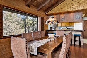 A wooden interior with a dining table, chairs, a kitchen area, and a large window with a view of trees. - 2600-the Tree Top Cabin (Big Bear Lake)