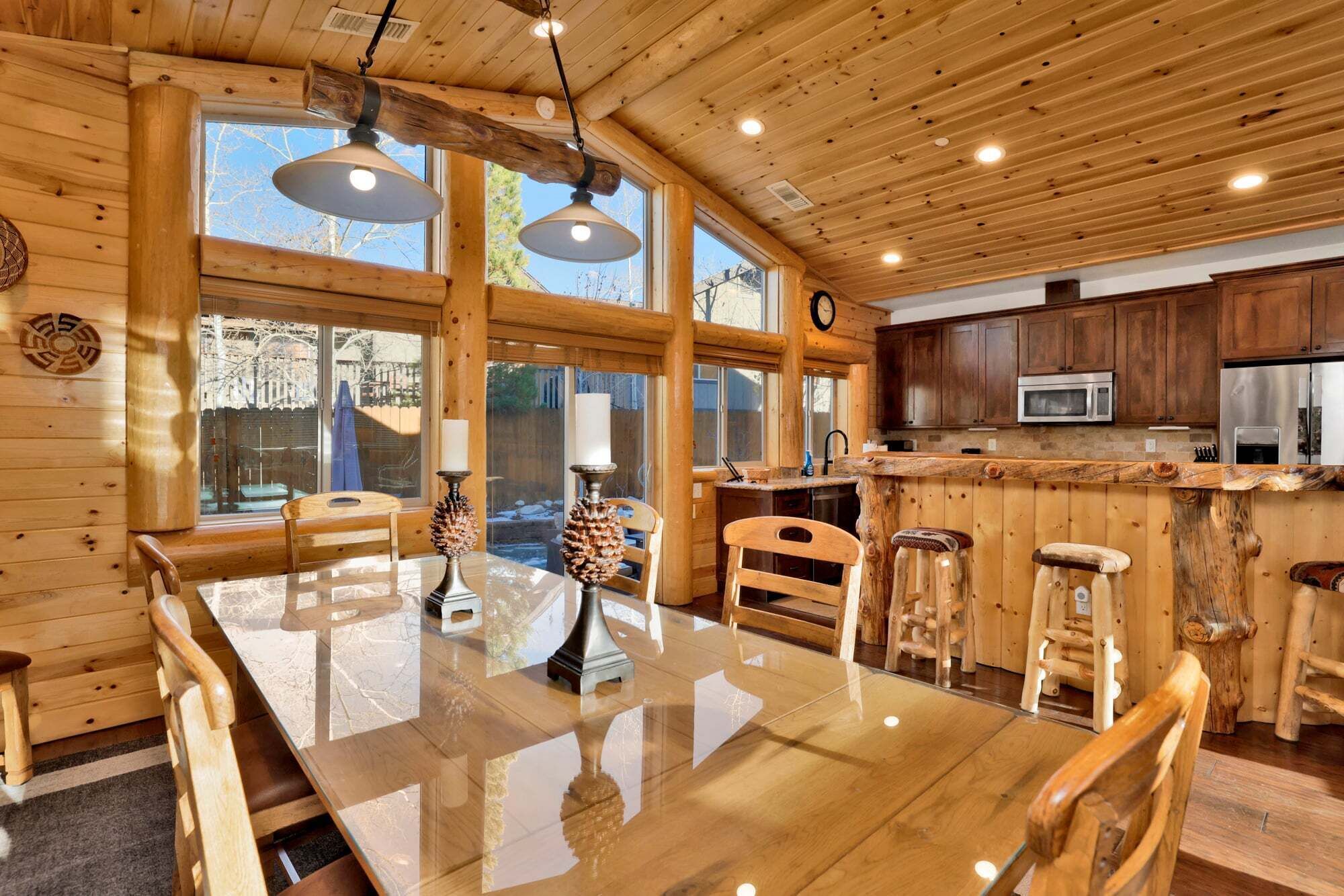 A wooden interior with a dining table, chairs, bar stools, a kitchen area, and large windows.