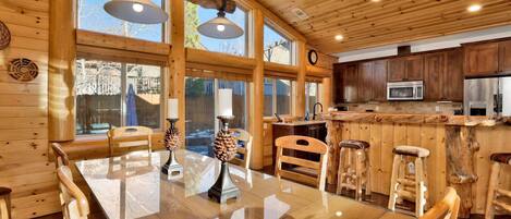 A wooden interior with a dining table, chairs, bar stools, a kitchen area, and large windows.