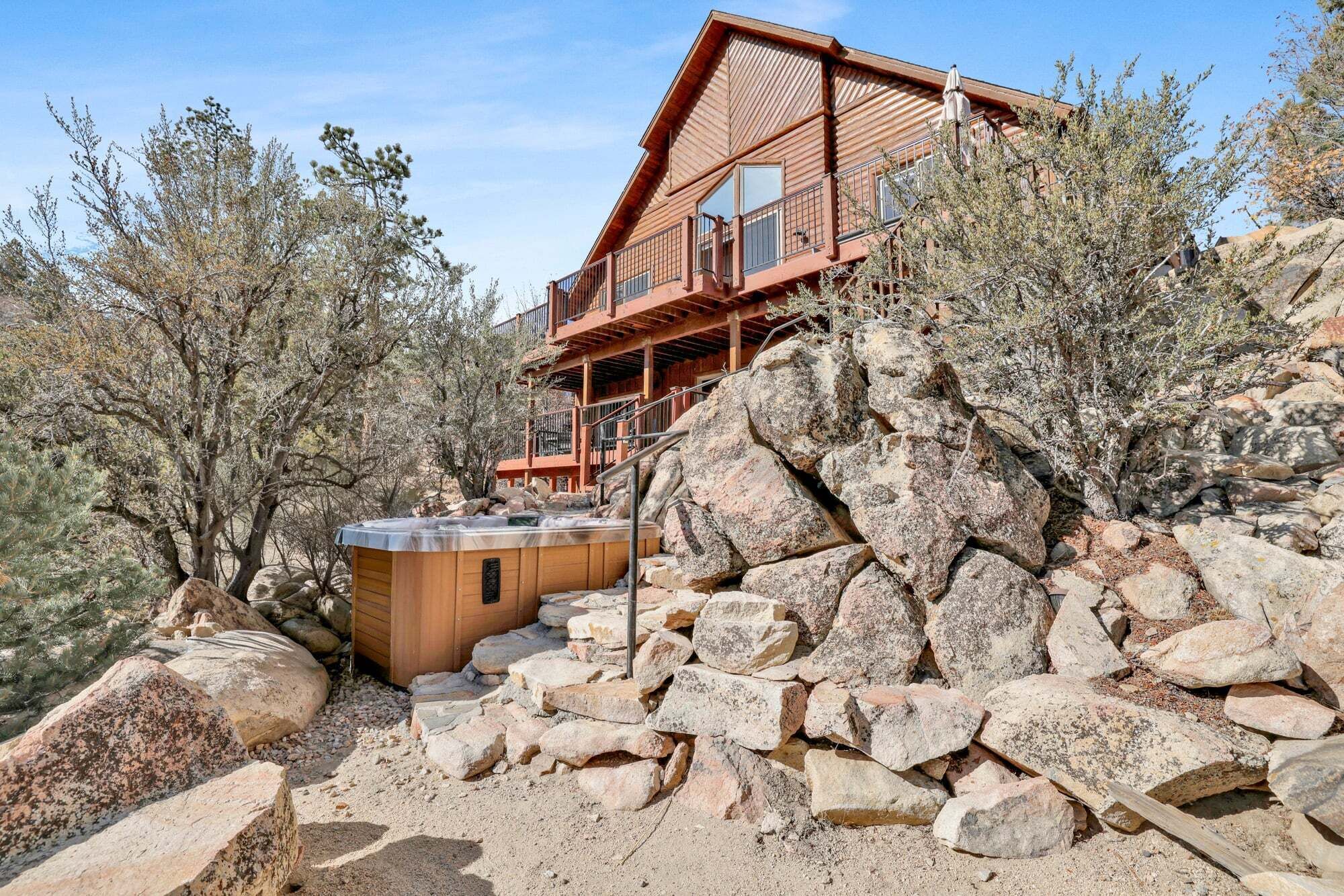 Una cabaña de madera con jacuzzi, rodeada de grandes rocas y vegetación escasa.