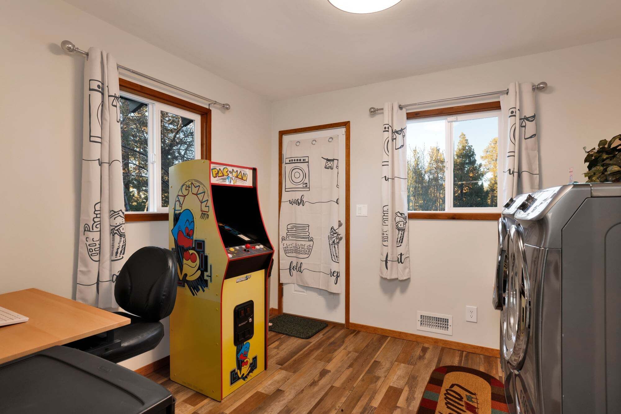 A room with a washing machine, a Pac-Man arcade machine, a desk with a chair, and two windows with curtains.