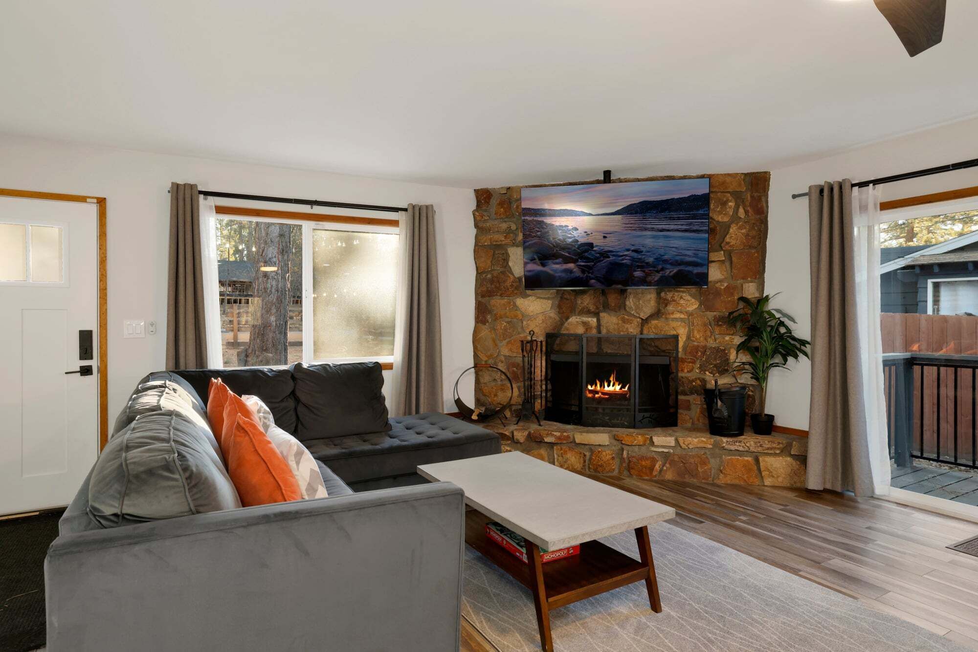 A living room with a stone fireplace, a large TV mounted above it, a grey sofa with orange cushions, and a view of the outdoors through a sliding glass door.