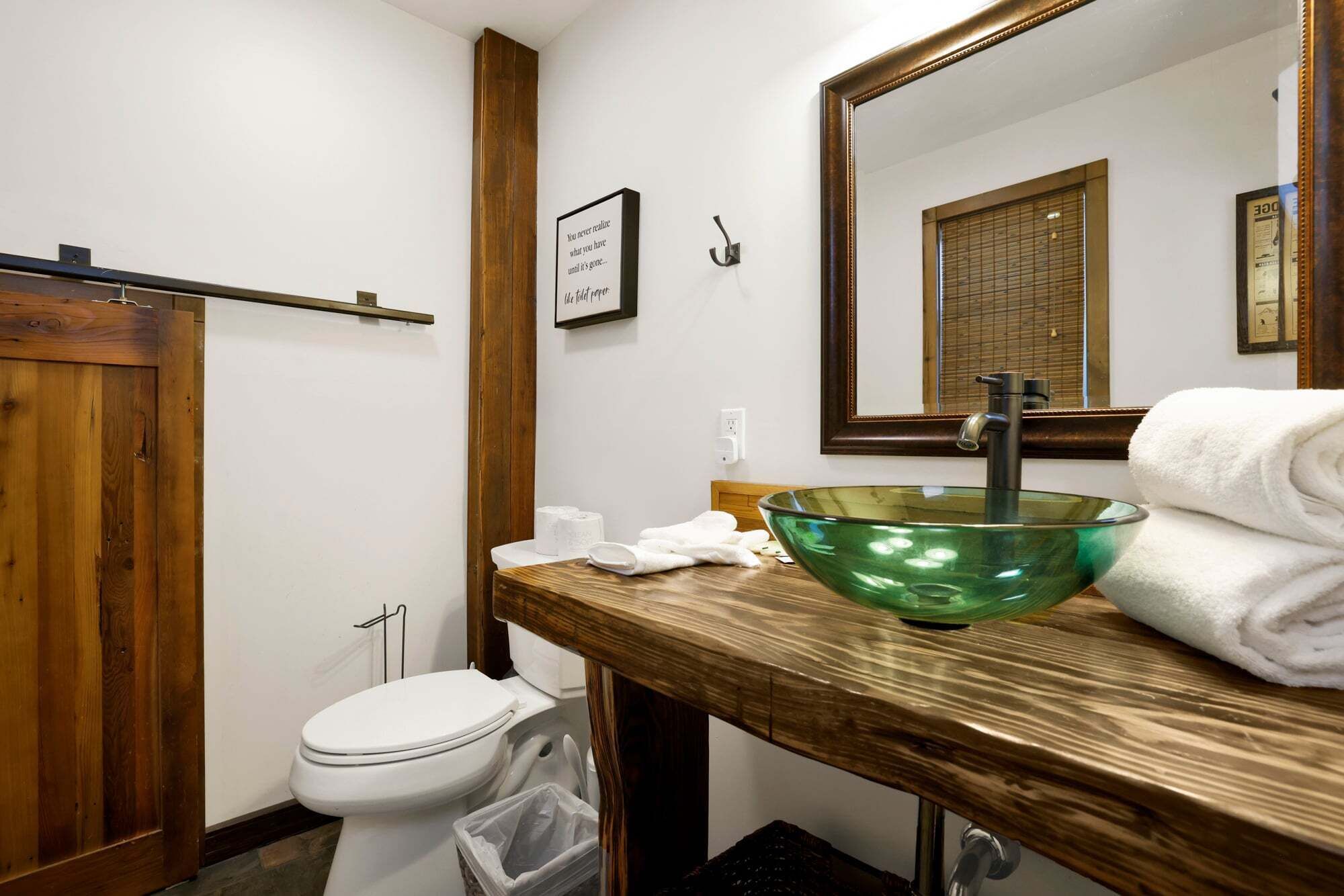 A bathroom with a wooden vanity, a green glass sink, a white toilet, and a mirror with a wooden frame.