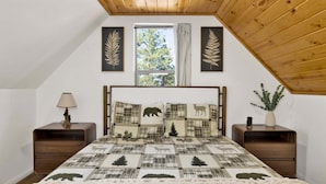 A bedroom with a sloped ceiling, a bed with a forest-themed quilt, two nightstands, a lamp, and a window with curtains. - 2613-shiloh Woods Cabin (Big Bear Lake)