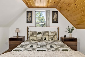 A bedroom with a sloped ceiling, a bed with a quilted headboard, two nightstands, a window with curtains, and a small plant on the nightstand. - 2613-shiloh Woods Cabin (Big Bear Lake)