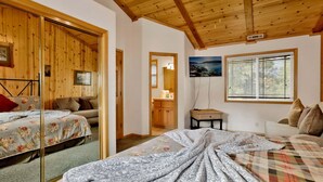 Une chambre avec un plafond en bois, un lit recouvert d’une literie à motifs floraux, une commode, une table de chevet et une fenêtre avec des stores.